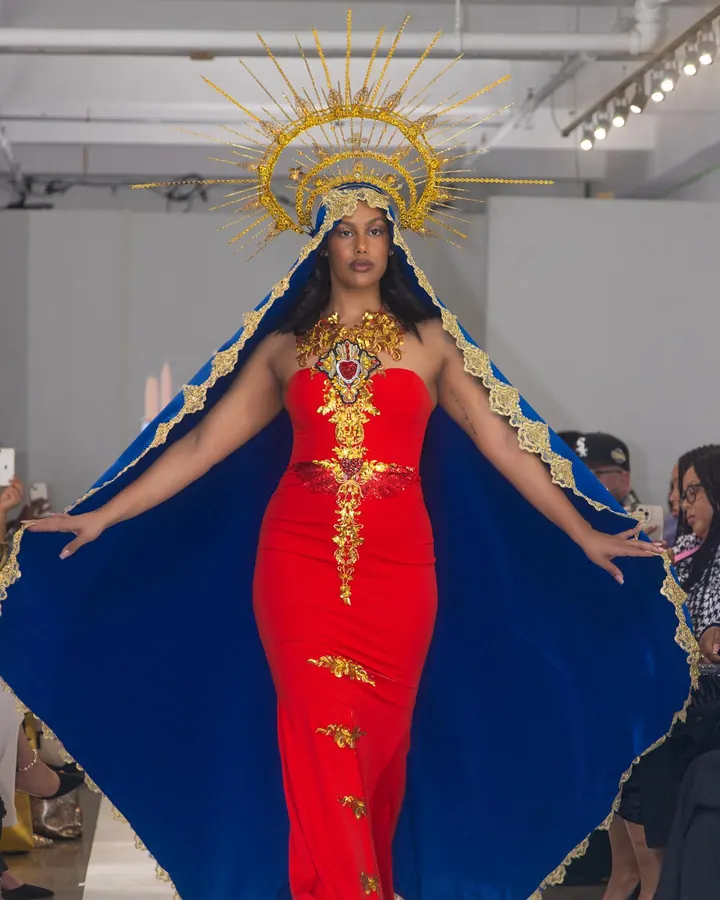 Model in a red gown, blue cape and gold halo with arms spread on the runway