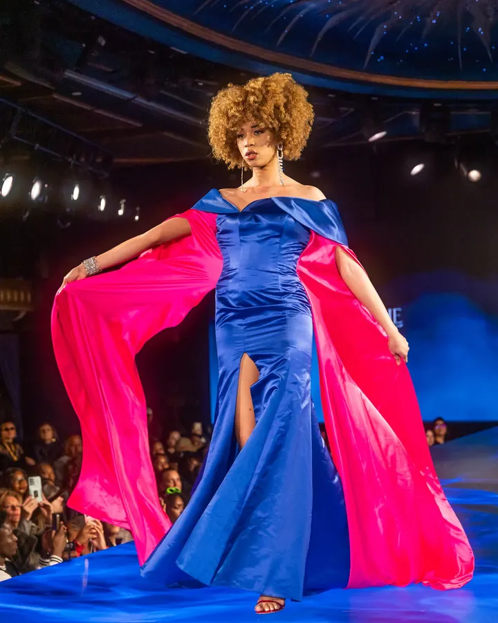 Model in a blue satin gown opening a pink cape on the runway